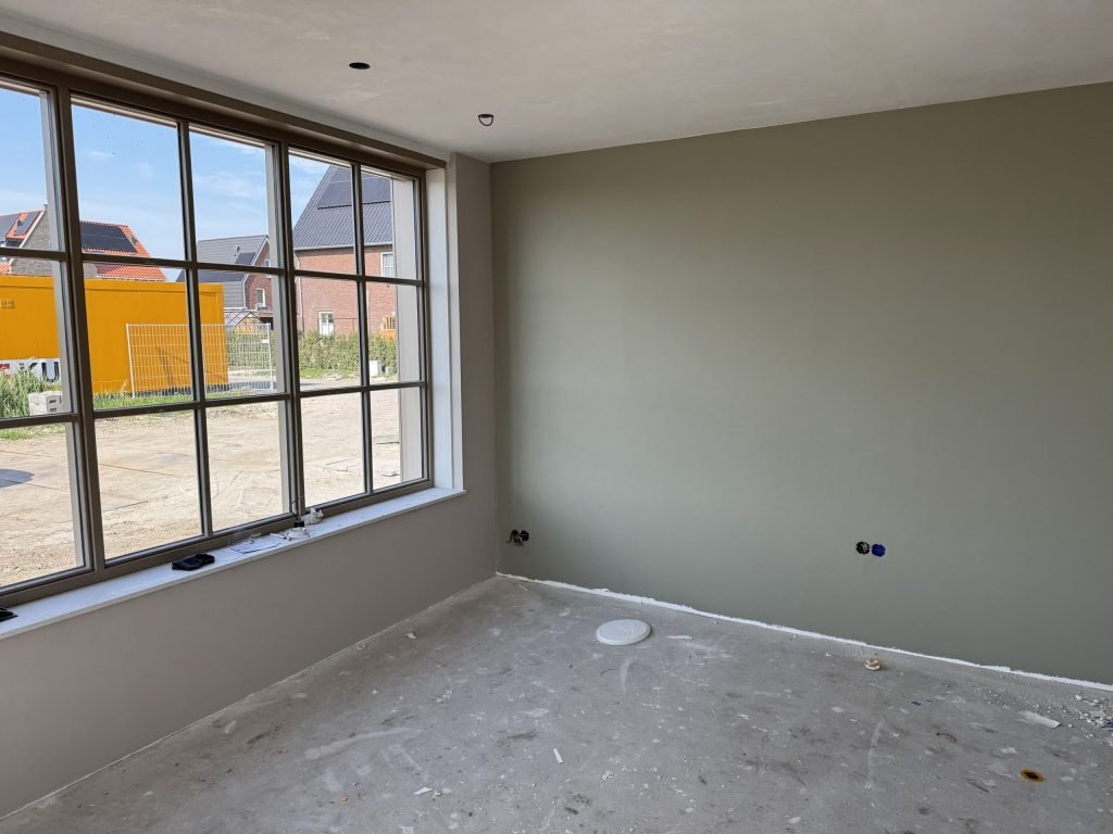 Kamer met een egaal gesausde wand in grijsgroen en een groot raam met zicht op nieuwbouw