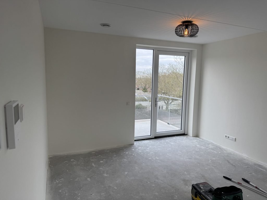 Lege nieuwbouwkamer met lichte wanden, balkondeuren en een kale vloer kort na de oplevering